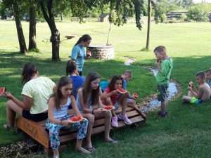Táborozó gyerekek dinnyét esznek a tanyaház előtt a Magyarkertben